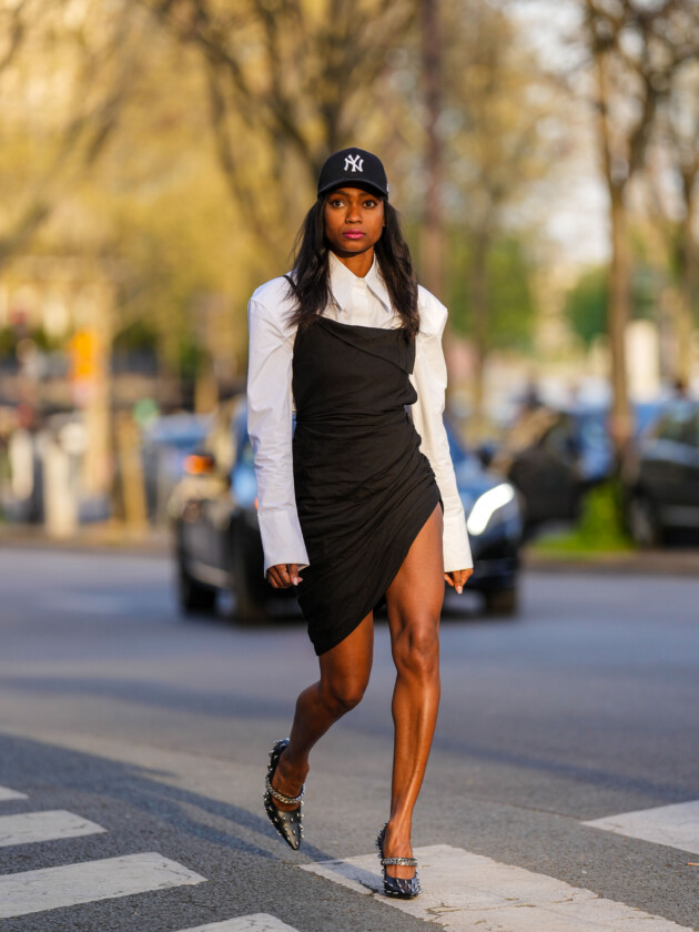 7 ideias para atualizar o slip dress neste inverno Mulher usa slip dress preto com camisa social branca por baixo e complementa o look com salto alto e boné