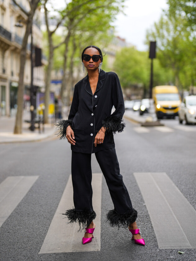 PARIS, FRANCE - MAY 05: Emilie Joseph @in_fashionwetrust wears silver rhinestones Isabel Marant earrings, black sunglasses, a black ribbed buttoned long shirt with black feathers on the wrists, daily sleeper matching pyjama set pants with black feathers on the ankle, a silver ring, fuchsia pointed pink Nicholas Kirkwood mules shoes with rhinestones detail, on May 05, 2021 in Paris, France. (Photo by Edward Berthelot/Getty Images) A tendência dos conjuntos femininos: Mulher usa conjunto preto de pijamas com plumas nas barras no street style