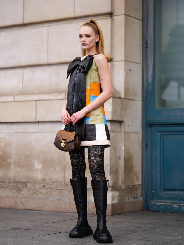 PARIS, FRANCE - MARCH 06: Emma Lair wears a blue / orange / black / white / green shiny leather yoke sleeveless / short dress with a black shiny varnished leather large knot embroidered on the neck from Louis Vuitton, a brown LV monogram print pattern in coated canvas handbag from Louis Vuitton, black mesh / fishnet with embroidered lace flower pattern tight from Louis Vuitton, gold and diamonds rings, black shiny leather platform soles knees boots / high boots from Louis Vuitton , outside Louis Vuitton, during Paris Fashion Week - Womenswear Fall Winter 2023 2024, on March 06, 2023 in Paris, France. (Photo by Edward Berthelot/Getty Images) Como atualizar o look com meia: adicione textura com uma meia-calça de renda