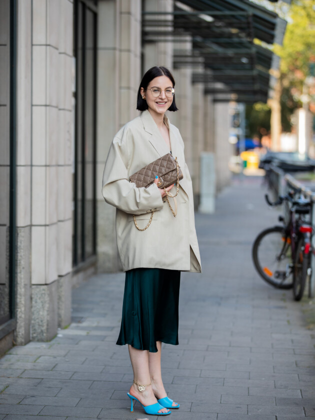 7 ideias para atualizar o slip dress neste inverno Combine o slip dress com blazer. Aqui, mulher usa blazer bege largo e combina com vestido acetinado azul